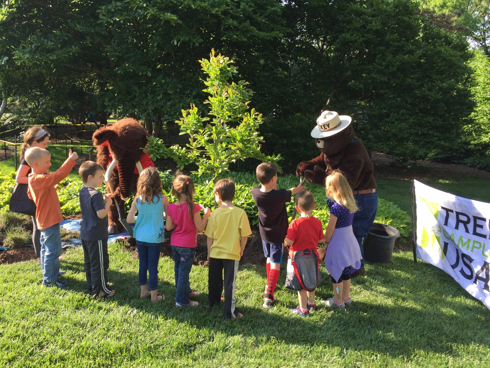 Children gather around newly planted tree and Smokey bear.