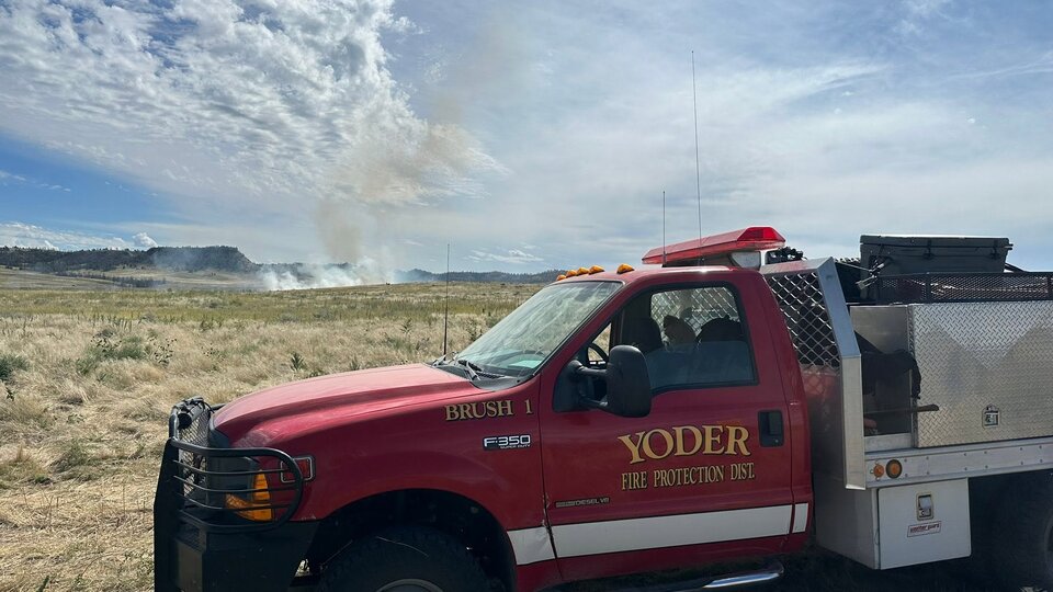 Red fire truck parked in dry grassland under a partly cloudy sky, with smoke rising in the distance.